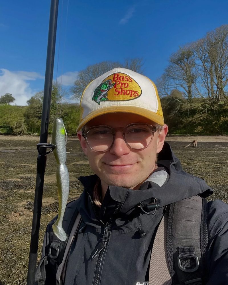 Lyle in a trucker cap and glasses, smiling, with a fishing rod over his shoulder